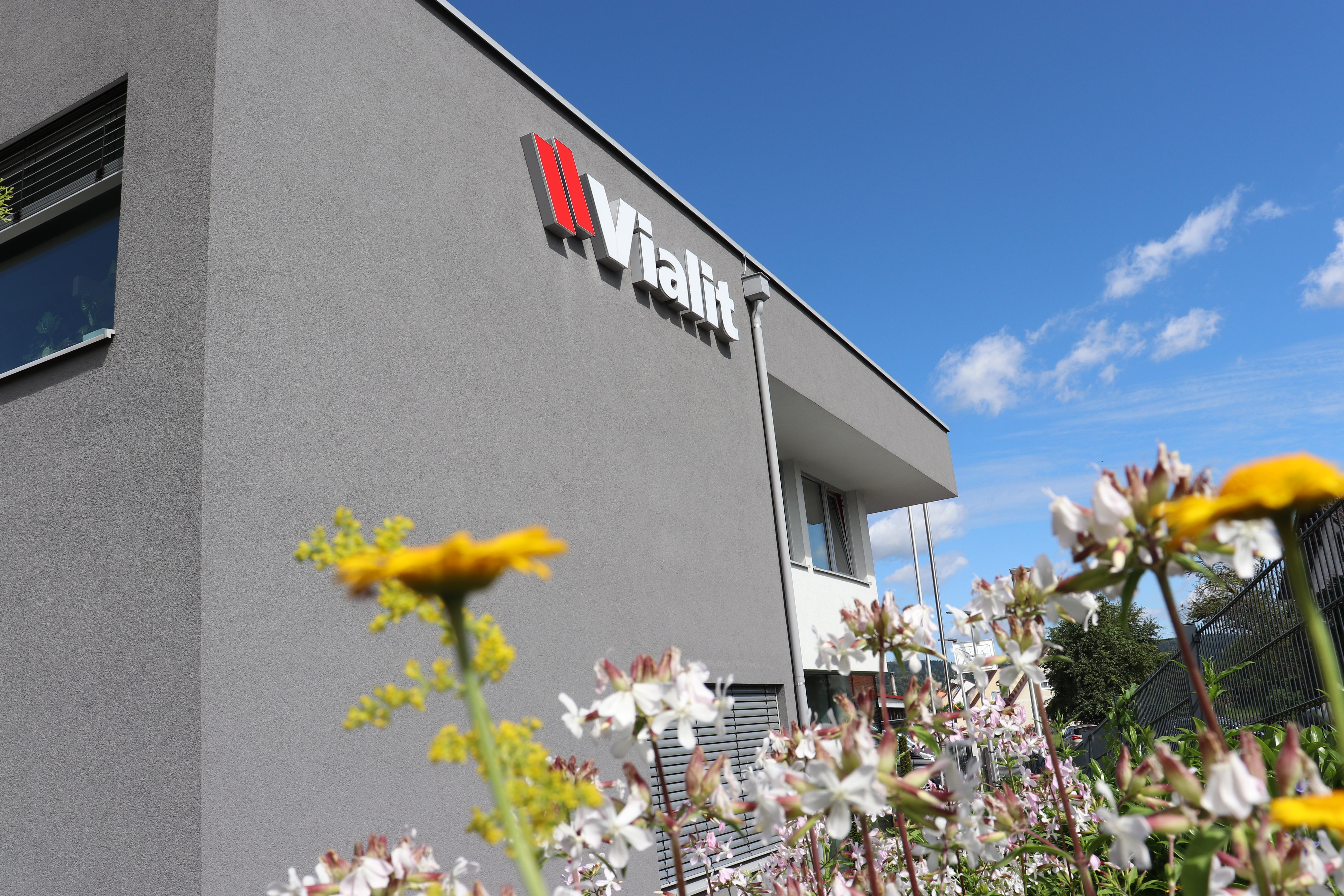 Viralit Firmengebäude mit blühenden Blumen im Vordergrund, modernes Design und klare Linien, unter blauem Himmel.