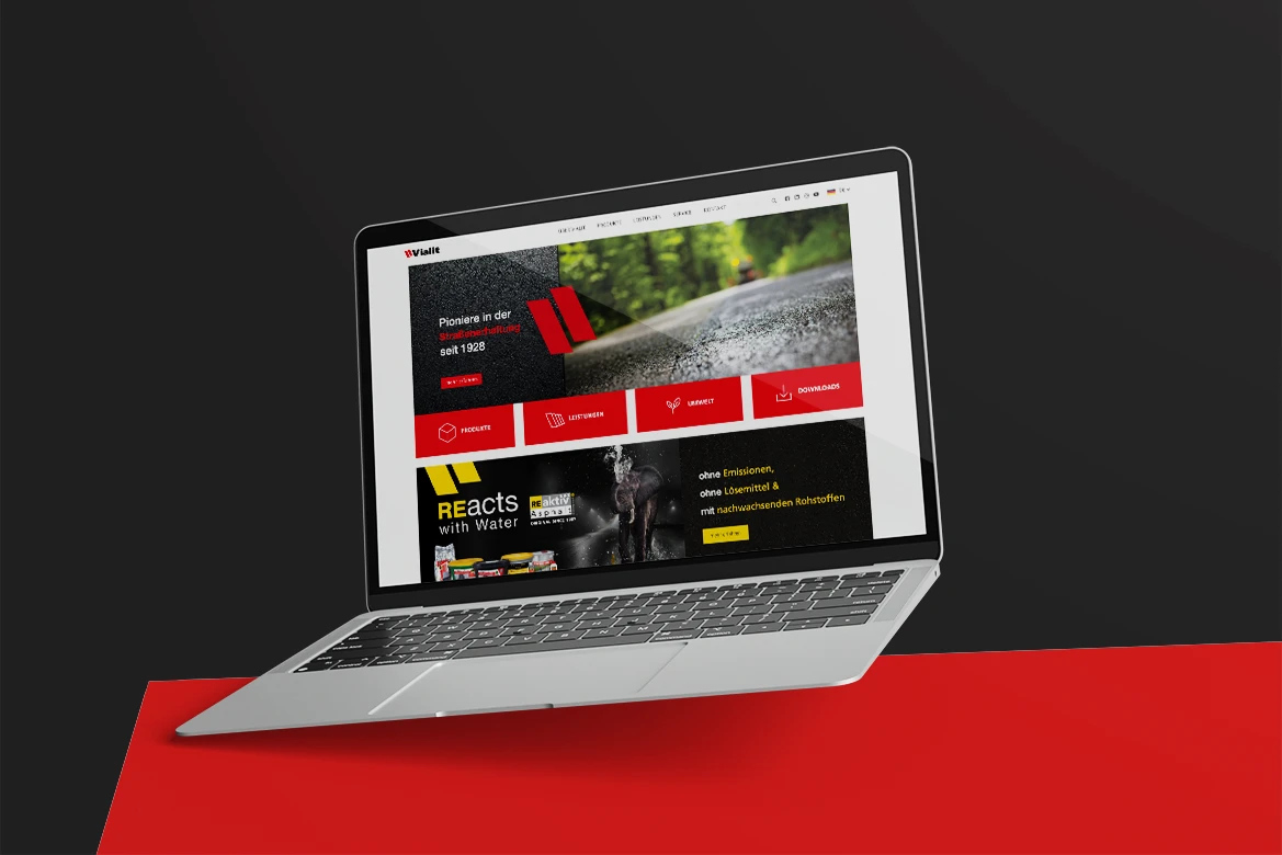 Laptop mit der Vialit Website, die Produkte und Dienstleistungen im Bereich Straßenbau präsentiert, auf rotem Hintergrund.
