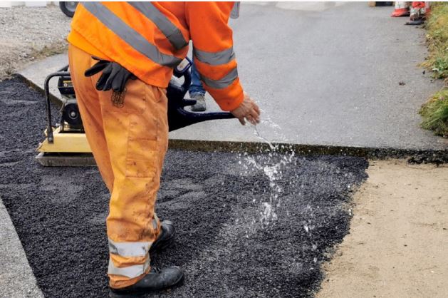Arbeiter gießt Wasser auf frischen Asphalt während der Verarbeitung, um die Qualität zu verbessern. Sicherheitskleidung und Ausrüstung sind sichtbar.